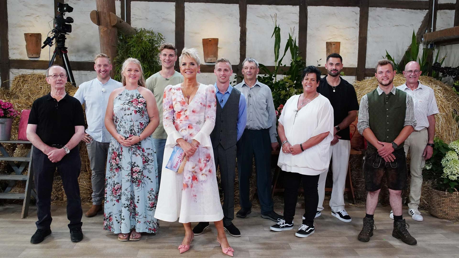 Auf dem Bild zur Sendung Bauer sucht Frau 2025 stehen die Moderatorin Inka Bause und die Teilnehmer:innen Walter, Daniel, Insa, Friedrich, Christopher, Johann, Simone, Basti, Michi und Herbert in einem rustikal dekorierten Studio mit Strohballen und Blumen.