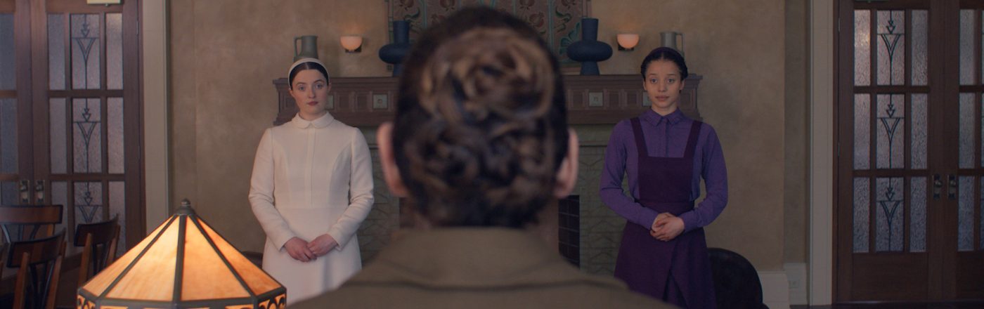 Auf dem Bild in dem Artikel zur neuen Serie „The Testaments“ stehen zwei junge Frauen nebeneinander in einem Raum, während eine Person mit gebundenem Haar im Vordergrund sitzt.