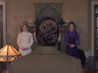 Auf dem Bild in dem Artikel zur neuen Serie „The Testaments“ stehen zwei junge Frauen nebeneinander in einem Raum, während eine Person mit gebundenem Haar im Vordergrund sitzt.