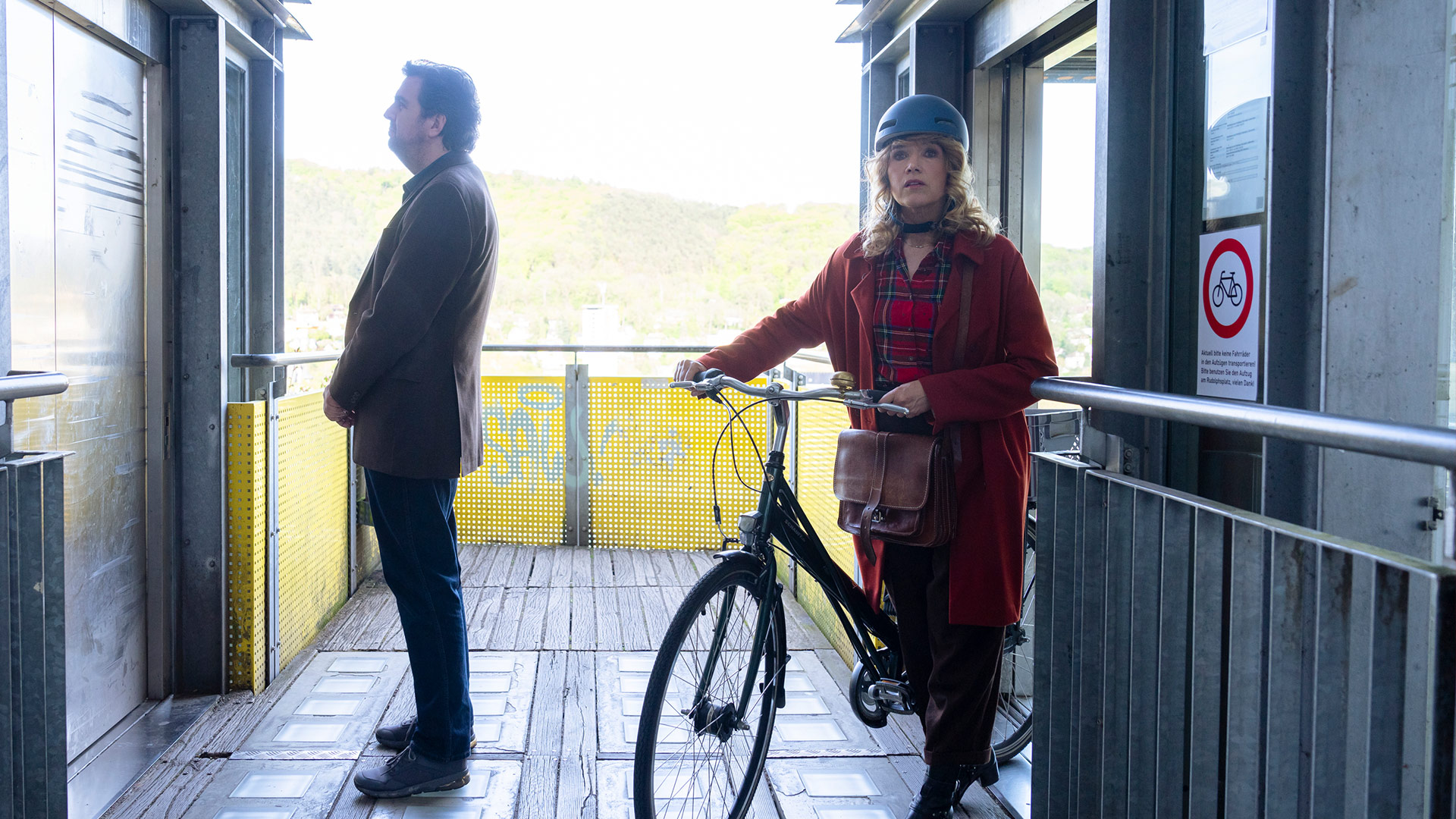 Auf dem Bild in dem Artikel zu Perfekt zusammen ist eine Szene aus der Prime-Video-Serie Perfekt verpasst zu sehen. Eine Frau und ein Mann stehen auf einer Plattform vor einem Aufzug. Die Frau trägt einen roten Mantel, einen Fahrradhelm und hält ein Fahrrad, während der Mann seitlich steht und ruhig wartet.