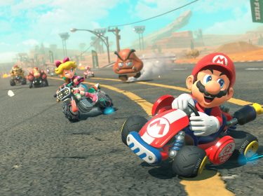Auf diesem Bild zu Mario Kart World Tipps driftet Mario fröhlich mit seinem roten Kart durch eine Wüstenlandschaft. Hinter ihm fährt Peach auf einem Motorrad und zwinkert in die Kamera, während weitere Fahrer und ein Goomba die Szene beleben. Die Grafik ist detailreich und dynamisch, was die rasante Rennatmosphäre perfekt einfängt.