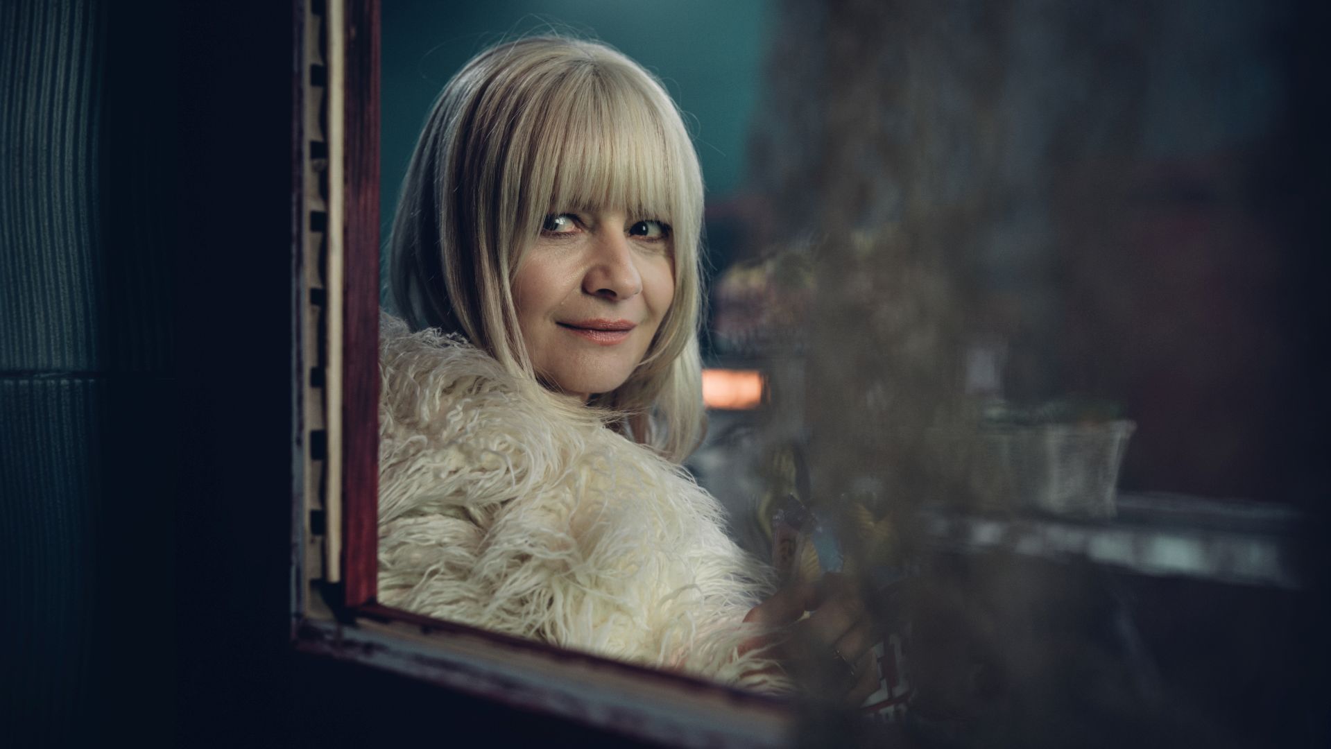 Małgorzata Kożuchowska in Aniela: Eine blonde Frau mit weißem Pelzmantel betrachtet sich lächelnd in einem Spiegel.