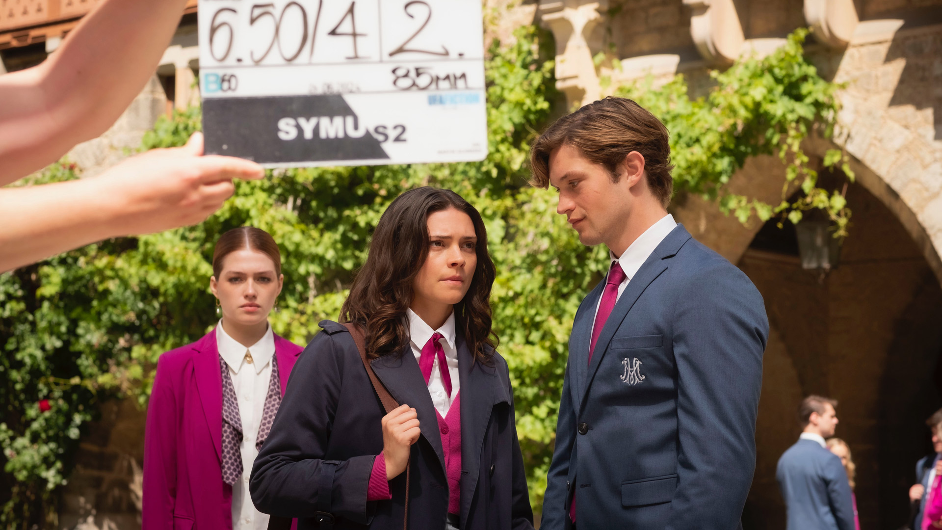 Auf diesem Making-of-Bild zu Maxton Hall Staffel 3 sind Harriet Herbig-Matten und Damian Hardung während der Dreharbeiten zu sehen. Harriet steht im Vordergrund mit ernster Miene, trägt eine Schuluniform mit pinker Weste und dunklem Mantel. Damian, im blauen Sakko mit Schullogo, schaut sie intensiv an. Im Hintergrund hält jemand eine Klappe in die Szene, was den Moment als Drehaufnahme kennzeichnet. Die Stimmung wirkt angespannt und konzentriert.