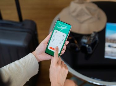 Eine Frau hält ein Smartphone in der Hand und nutzt eine der Reiseplanungs-Apps, um einen Flug von London nach Sydney zu buchen. Im Hintergrund stehen ein schwarzer Koffer, ein Sonnenhut und eine Kamera auf einem runden Tisch, daneben liegt ein Reisemagazin. Die Szene vermittelt Vorfreude und Reiselust.