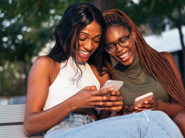 Zwei junge Frauen sitzen auf einer Bank, schauen auf ihre Smartphones und lachen.