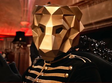 Symbolbild aus The 50 Staffel 3: Der Spielleiter ist als geheimnisvoller Löwe dargestellt, mit einer goldenen, geometrischen Maske und einer schwarzen Paradeuniform mit goldenen Applikationen. Die Szene spielt in einem prunkvoll beleuchteten Raum mit rotem Lichtakzent. Die Figur wirkt dominant und rätselhaft – passend zur Rolle des Spielleiters in The 50 Staffel 3.