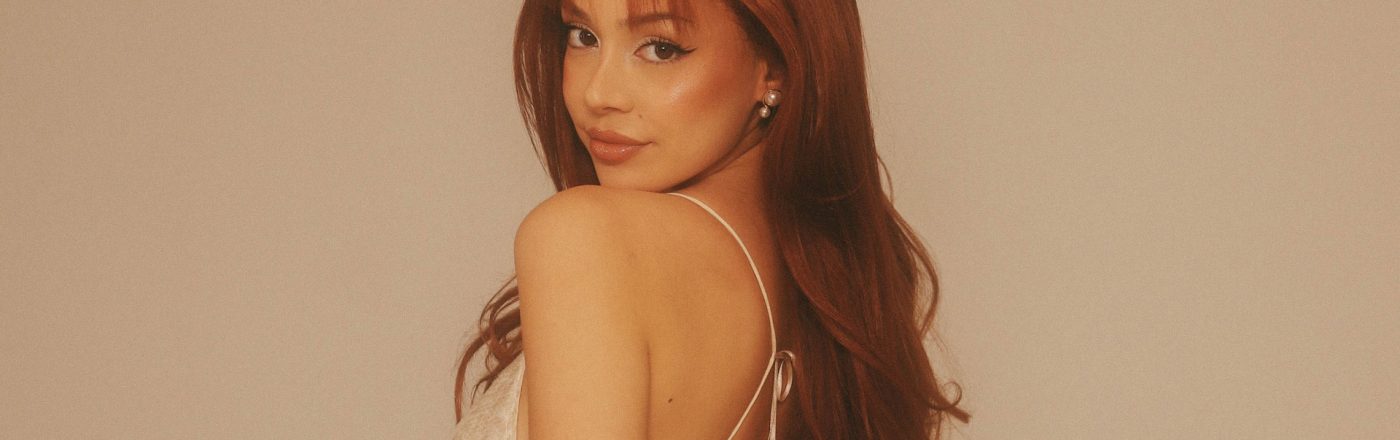 Für den Artikel über ayliva tickets zeigt das Bild Sängerin Ayliva mit rotbraunen, offenen Haaren und einem hellen, schimmernden Top mit dünnen Trägern. Sie blickt über die Schulter zur Kamera, vor einem neutralen, beigen Hintergrund.