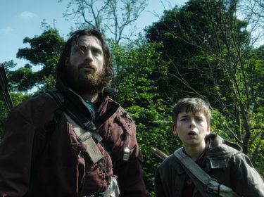 In dieser Szene aus 28 Years Later stehen Jamie, gespielt von Aaron Taylor-Johnson, und sein Sohn Spike, gespielt von Alfie Williams, inmitten eines dichten Waldes. Beide blicken alarmiert nach oben, mit ernsten, angespannten Gesichtern. Jamie trägt einen Köcher mit Pfeilen auf dem Rücken