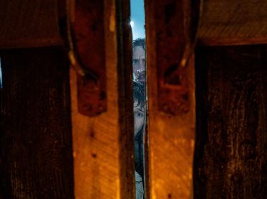 Das Bild aus der Filmkritik zu 28 Years Later zeigt eine intensive Szene mit Jamie (Aaron Taylor-Johnson) und Spike (Alfie Williams), die durch einen schmalen Spalt in einer massiven Holztür blicken. Ihre Gesichter wirken angespannt und vorsichtig, während nur wenig Licht ins Bild fällt – die bedrohliche Stimmung unterstreicht das post-apokalyptische Setting eindrucksvoll.