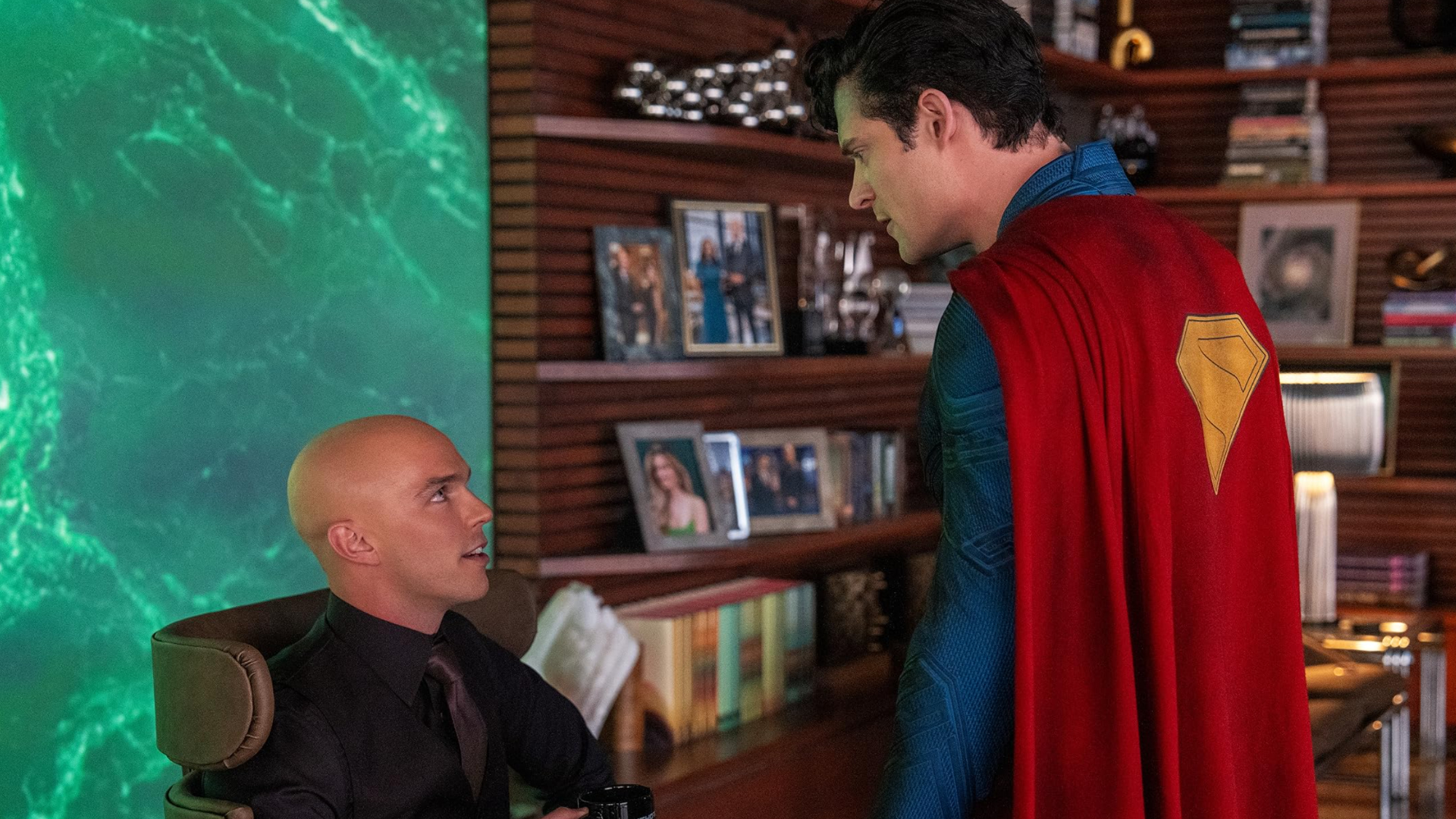 Nicholas Hoult und David Corenswet in Superman: in Mann mit Glatze sitzt auf einem Ledersessel, hält eine Kaffetasse in der Hand und blick hoch zu dem vor ihm stehenden Superman.