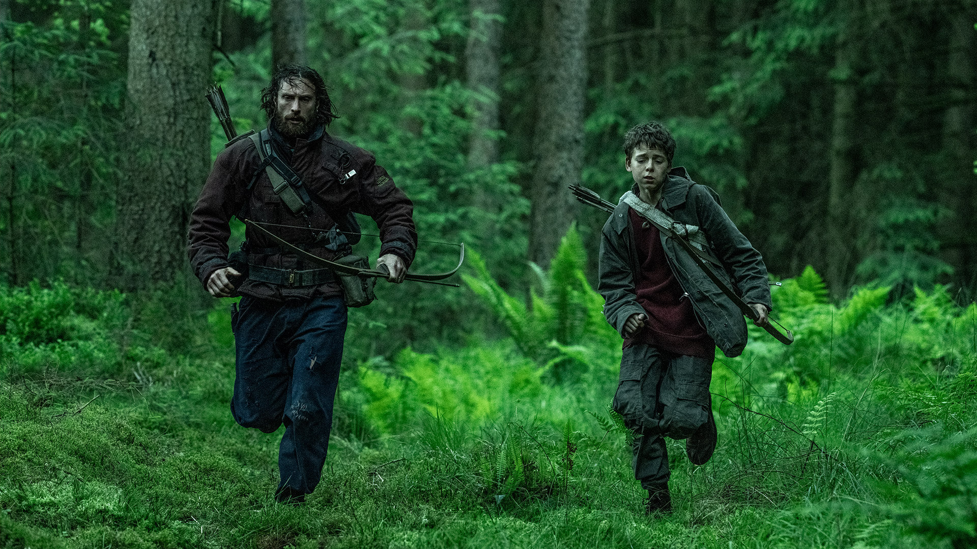 Auf dem Bild zur 28 Years Later-Kritik sind Jamie, gespielt von Aaron Taylor-Johnson, und Spike, dargestellt von Alfie Williams, in einer spannungsgeladenen Szene im dichten Wald zu sehen. Beide tragen dunkle Kleidung und Bögen, während sie durch das grüne Unterholz rennen. Die Atmosphäre wirkt düster und bedrohlich, passend zum post-apokalyptischen Setting des Films.