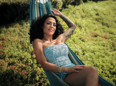 Auf dem Bild zu Make Love Fake Love Staffel 4 liegt Elena Miras entspannt in einer Hängematte. Sie trägt ein hellblaues, schulterfreies Outfit mit Blumenstickerei, ihr lockiges Haar fällt über die Schultern. Der Hintergrund zeigt eine grüne Wiese mit kleinen roten Blüten.