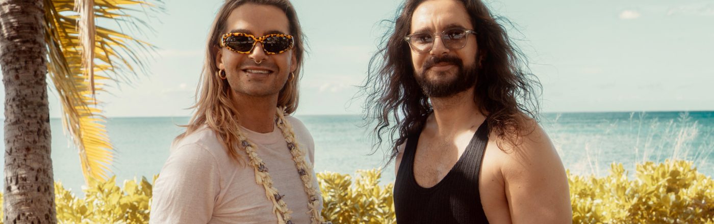 Auf dem Bild zur möglichen Fortsetzung Kaulitz & Kaulitz Staffel 3 stehen Bill und Tom Kaulitz entspannt vor tropischer Kulisse mit Meerblick. Bill trägt eine auffällige Sonnenbrille, ein weißes Shirt und eine Blumenkette, während Tom mit langem Haar und schwarzem Tanktop lässig wirkt. Die Stimmung ist sommerlich und gelöst – ein symbolischer Ausblick auf neue Folgen.