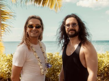 Auf dem Bild zur möglichen Fortsetzung Kaulitz & Kaulitz Staffel 3 stehen Bill und Tom Kaulitz entspannt vor tropischer Kulisse mit Meerblick. Bill trägt eine auffällige Sonnenbrille, ein weißes Shirt und eine Blumenkette, während Tom mit langem Haar und schwarzem Tanktop lässig wirkt. Die Stimmung ist sommerlich und gelöst – ein symbolischer Ausblick auf neue Folgen.