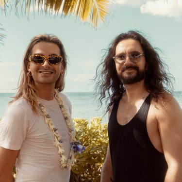 Auf dem Bild zur möglichen Fortsetzung Kaulitz & Kaulitz Staffel 3 stehen Bill und Tom Kaulitz entspannt vor tropischer Kulisse mit Meerblick. Bill trägt eine auffällige Sonnenbrille, ein weißes Shirt und eine Blumenkette, während Tom mit langem Haar und schwarzem Tanktop lässig wirkt. Die Stimmung ist sommerlich und gelöst – ein symbolischer Ausblick auf neue Folgen.