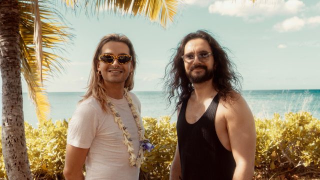Auf dem Bild zur möglichen Fortsetzung Kaulitz & Kaulitz Staffel 3 stehen Bill und Tom Kaulitz entspannt vor tropischer Kulisse mit Meerblick. Bill trägt eine auffällige Sonnenbrille, ein weißes Shirt und eine Blumenkette, während Tom mit langem Haar und schwarzem Tanktop lässig wirkt. Die Stimmung ist sommerlich und gelöst – ein symbolischer Ausblick auf neue Folgen.