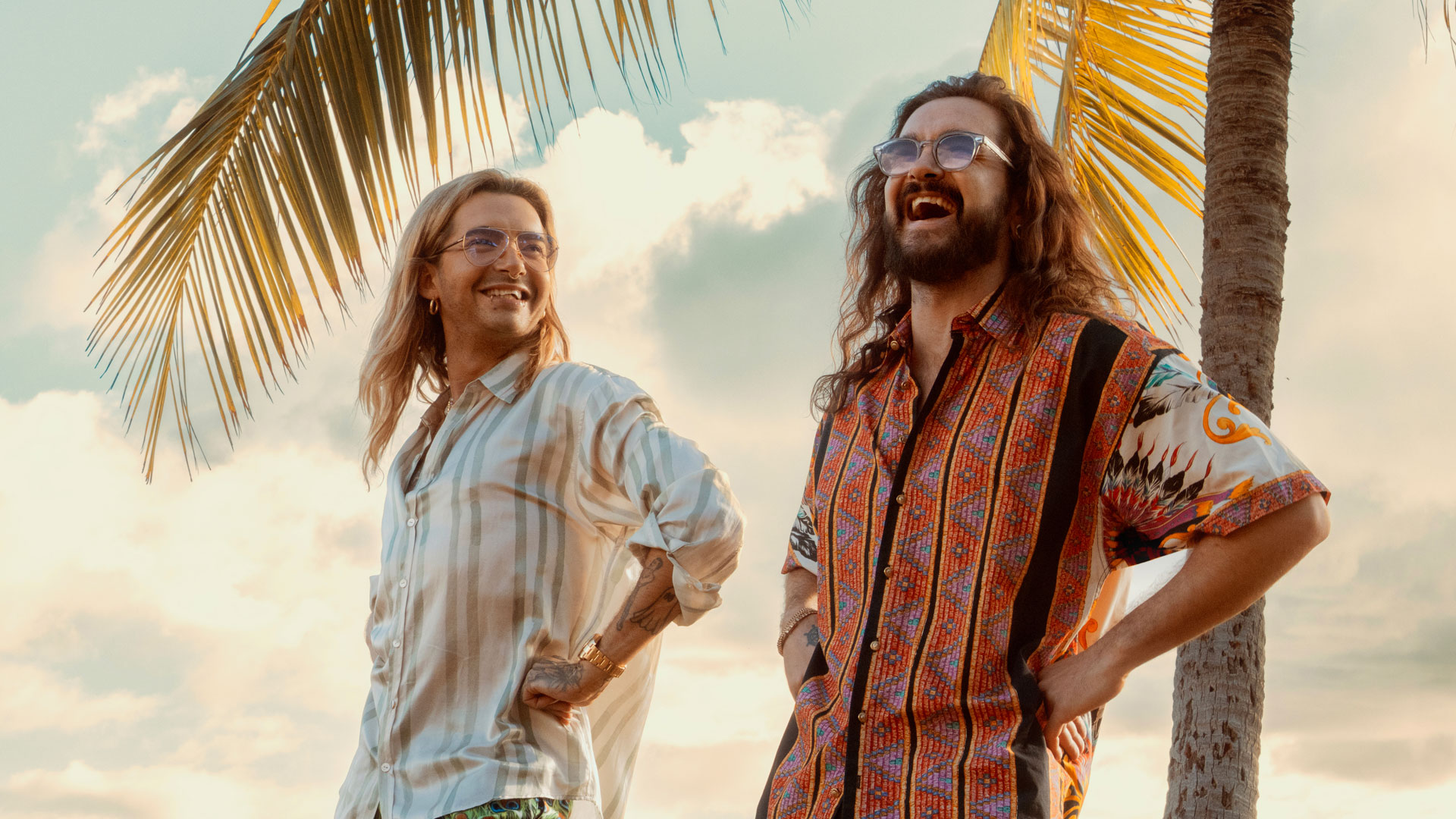 Auf dem symbolischen Bild zur Kaulitz & Kaulitz Staffel 3 stehen Bill und Tom Kaulitz entspannt unter Palmen im Sonnenuntergang. Bill trägt ein hell gestreiftes Hemd und blickt lächelnd zu seinem Bruder, der mit farbenfrohem Musterhemd herzhaft lacht. Die Stimmung ist unbeschwert und sommerlich – ein Hinweis auf mögliche neue Folgen.