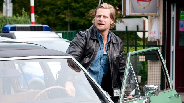 Auf dem Bild zur Serie Der letzte Bulle ist ein blonder Mann mit Bart zu sehen, der eine schwarze Lederjacke und ein offenes Jeanshemd trägt. Er steigt gerade aus einem mintgrünen Oldtimer mit geöffneter Fahrertür. Im Hintergrund steht ein Polizeiwagen.
