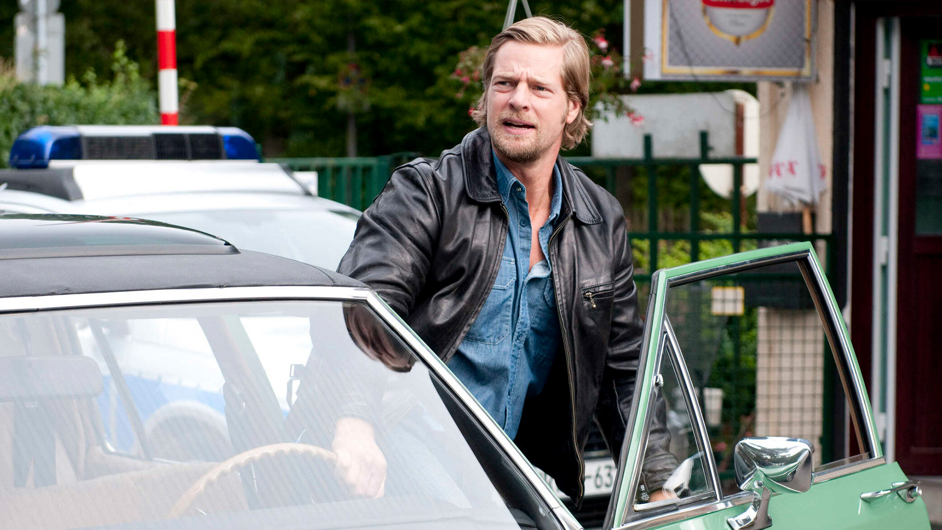 Auf dem Bild zur Serie Der letzte Bulle ist ein blonder Mann mit Bart zu sehen, der eine schwarze Lederjacke und ein offenes Jeanshemd trägt. Er steigt gerade aus einem mintgrünen Oldtimer mit geöffneter Fahrertür. Im Hintergrund steht ein Polizeiwagen.