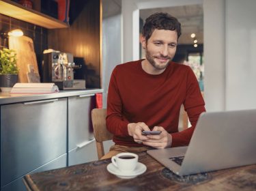 Vodafone E-Mail & Cloud im Netzwelt-Test: Mann in roten Pullover sitzt mit einem Kaffee vor seinem Laptop in einer modern eingerichteten Küche.