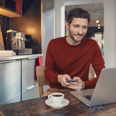 Vodafone E-Mail & Cloud im Netzwelt-Test: Mann in roten Pullover sitzt mit einem Kaffee vor seinem Laptop in einer modern eingerichteten Küche.