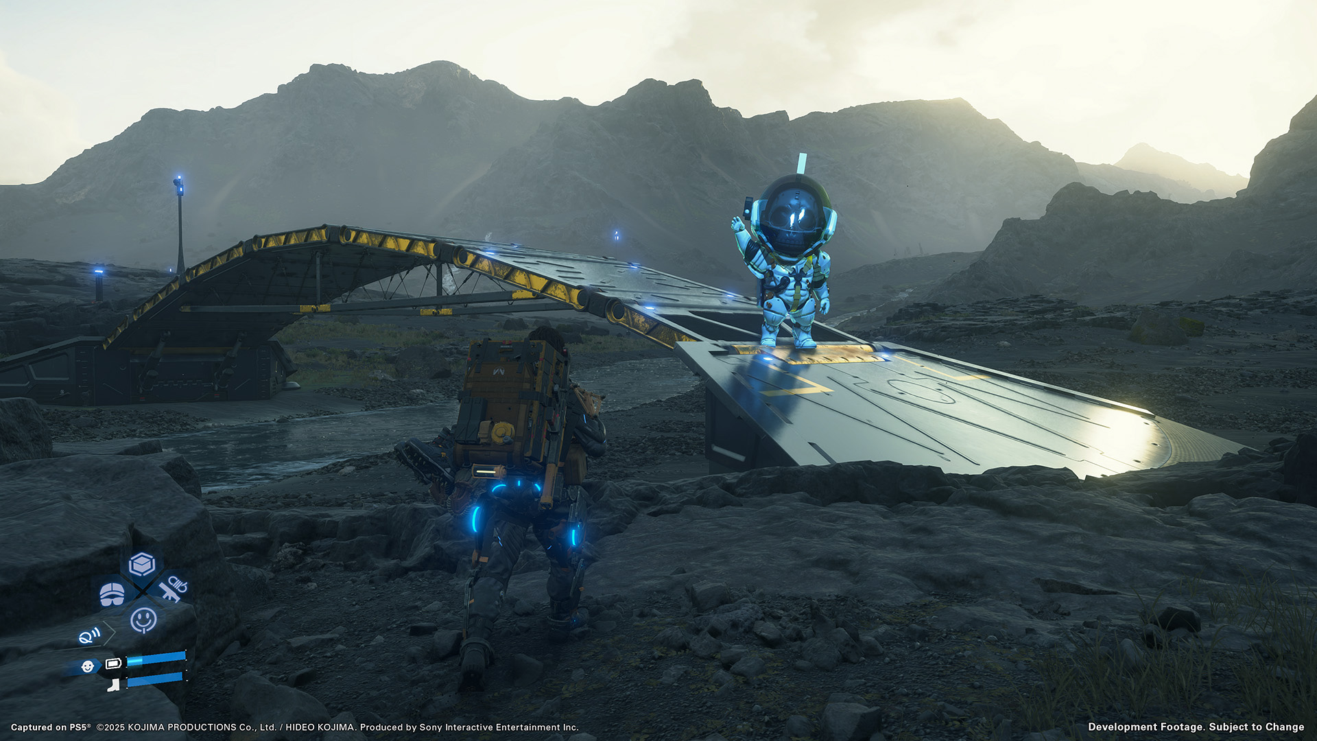 Auf diesem Screenshot aus Death Stranding 2: On the Beach nähert sich ein Bote mit großem Rucksack einer futuristischen Brücke in einer felsigen Landschaft. Am Brückenanfang steht ein leuchtend blauer, animierter Hologramm-Astronaut und winkt freundlich. Die Szene wirkt ruhig und technologisch fortschrittlich.