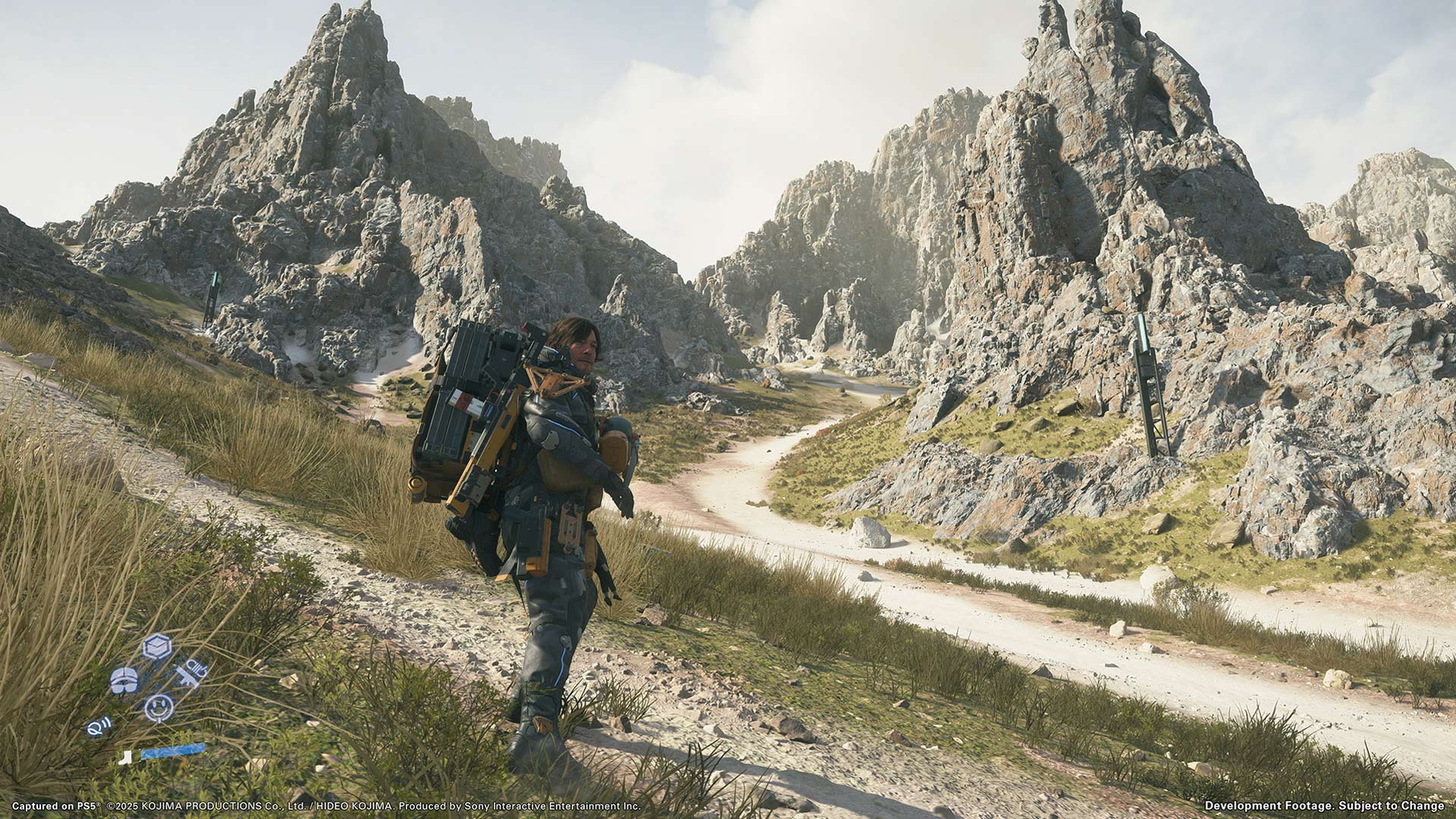 Im Bild aus Death Stranding 2 durchquert Sam Porter Bridges, gespielt von Norman Reedus, eine weitläufige Berglandschaft mit rauem Gelände und steilen Felsformationen. Er trägt schweres Gepäck auf dem Rücken und blickt entschlossen nach vorn. Die Szene symbolisiert die einsame Reise und die Herausforderungen innerhalb der Death Stranding 2 Story.