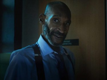 In den GigaTV-Highlights im Juli 2025 siehst du ein Bild aus Final Destination 6: Bloodlines: Tony Todd ist als William John Bludworth zu sehen. Er trägt ein hellblaues Hemd mit Krawatte und Hosenträgern und steht vor einer Tür in dunkler Umgebung.