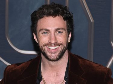 Auf diesem Bild ist Schauspieler Aaron Taylor-Johnson bei einem öffentlichen Auftritt zu sehen. Er trägt ein offenes, schwarzes Hemd unter einem braunen Samt-Sakko und mehrere goldene Ketten. Mit einem breiten Lächeln blickt er direkt in die Kamera. Das Bild lässt sich gut im Kontext aktueller Aaron Taylor-Johnson Filme verwenden.