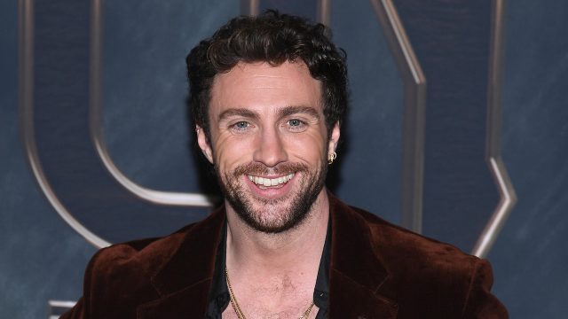 Auf diesem Bild ist Schauspieler Aaron Taylor-Johnson bei einem öffentlichen Auftritt zu sehen. Er trägt ein offenes, schwarzes Hemd unter einem braunen Samt-Sakko und mehrere goldene Ketten. Mit einem breiten Lächeln blickt er direkt in die Kamera. Das Bild lässt sich gut im Kontext aktueller Aaron Taylor-Johnson Filme verwenden.