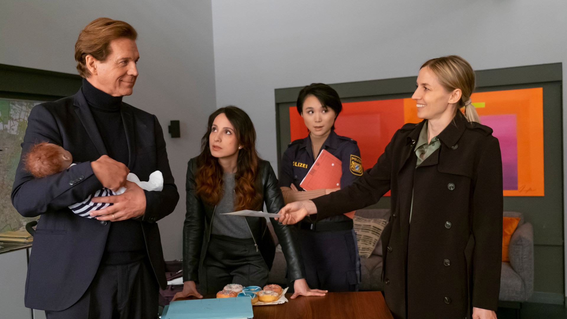 Das Artikelbild zu den Der Alte-Darsteller:innen zeigt Thomas Heinze als Hauptkommissar Caspar Bergmann, Stephanie Stumph als Kommissarin Annabell Lorenz, Yun Huang als IT-Spezialistin Julia Lulu Zhao und Annika Blendl als Richterin Eva Reinhardt (v.l.n.r.) zu sehen. Die Szene zeigt ein Gespräch im Büro, Heinze hält ein Baby, während Blendl ein Dokument überreicht.