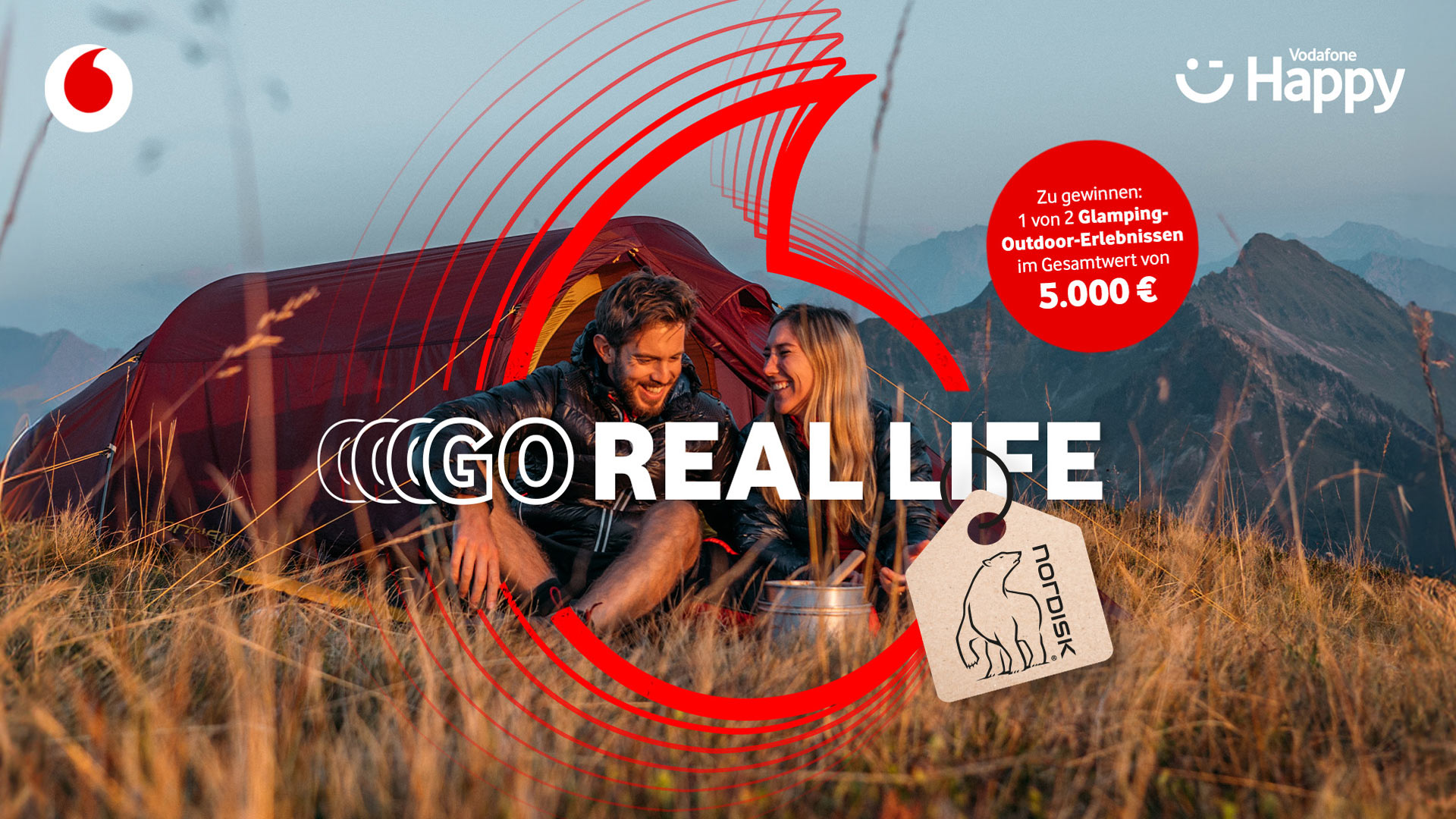 In dieser Szene der Vodafone-Kampagne „GO Real Life“ sitzt ein junges Paar lachend vor einem roten Nordisk-Zelt inmitten einer Berglandschaft. Die warme Abendstimmung unterstreicht das Naturerlebnis. Die „GO Real Life“-Werbung wirbt für ein Glamping-Gewinnspiel im Wert von 5.000 € und zeigt Naturverbundenheit und Lebensfreude.