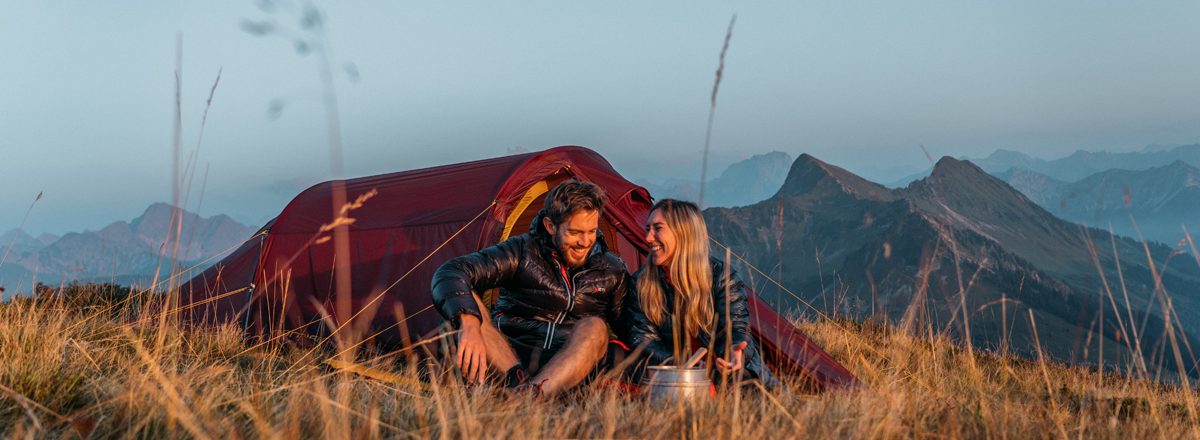 Auf diesem Bild der GO Real Life-Kampagne sitzen ein Mann und eine Frau entspannt im Gras vor einem roten Nordisk-Zelt. Die beiden lachen sich an und genießen die Aussicht auf eine Berglandschaft bei Sonnenaufgang oder Sonnenuntergang. Die Szene vermittelt Ruhe, Naturverbundenheit und echte Outdoor-Erlebnisse.