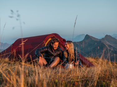 Auf diesem Bild der GO Real Life-Kampagne sitzen ein Mann und eine Frau entspannt im Gras vor einem roten Nordisk-Zelt. Die beiden lachen sich an und genießen die Aussicht auf eine Berglandschaft bei Sonnenaufgang oder Sonnenuntergang. Die Szene vermittelt Ruhe, Naturverbundenheit und echte Outdoor-Erlebnisse.