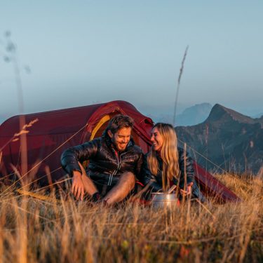 Auf diesem Bild der GO Real Life-Kampagne sitzen ein Mann und eine Frau entspannt im Gras vor einem roten Nordisk-Zelt. Die beiden lachen sich an und genießen die Aussicht auf eine Berglandschaft bei Sonnenaufgang oder Sonnenuntergang. Die Szene vermittelt Ruhe, Naturverbundenheit und echte Outdoor-Erlebnisse.