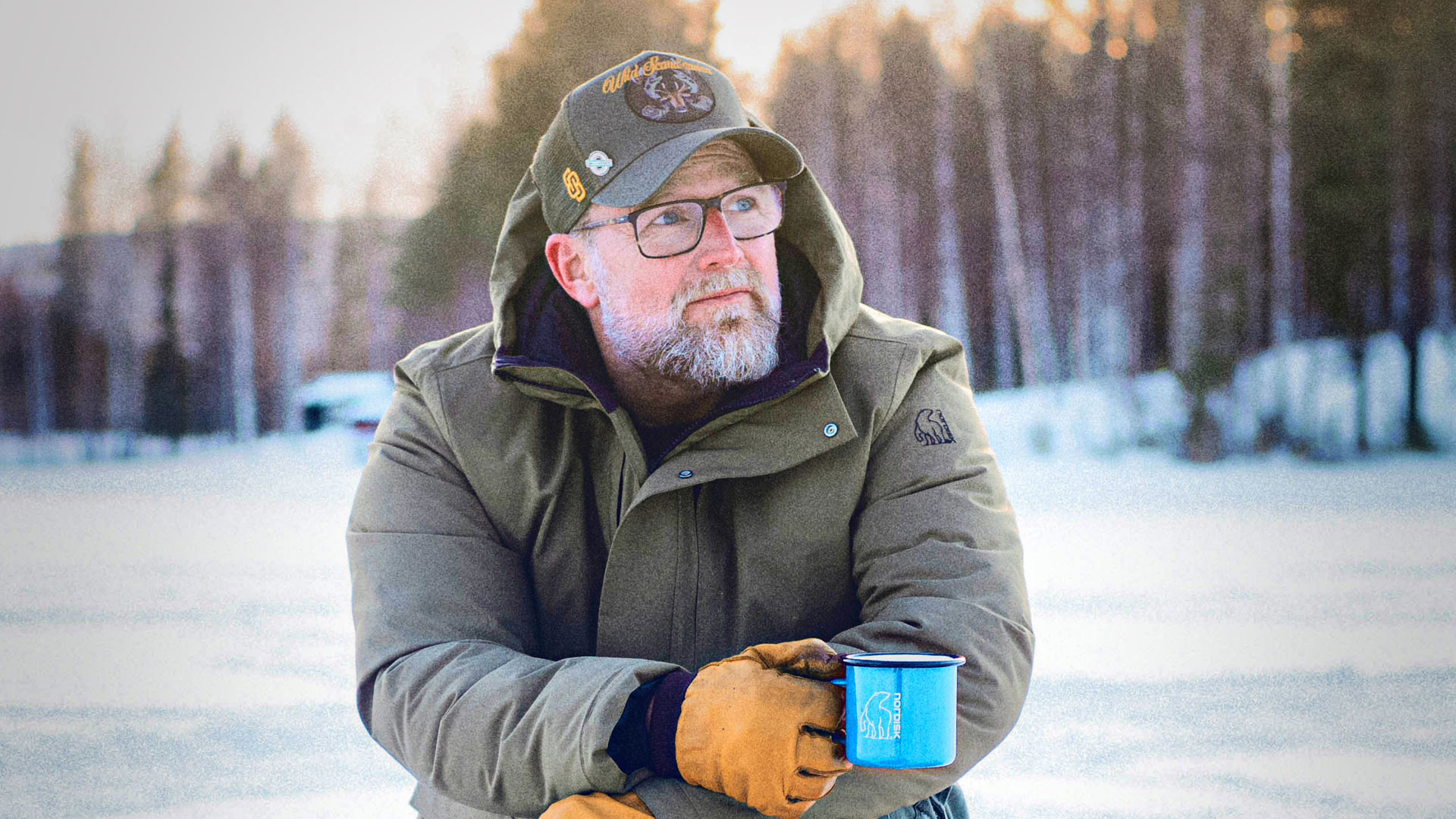 Auf dem Bild zur GO Real Life-Kampagne ist Brian Bojsen in winterlicher Umgebung zu sehen. Er trägt eine grüne Outdoorjacke mit Nordisk-Logo, eine Baseballkappe und Handschuhe. In der Hand hält er eine blaue Emaille-Tasse von Nordisk. Der Blick in die Ferne wirkt ruhig und reflektiert – die Szene strahlt Naturverbundenheit und Gelassenheit aus.