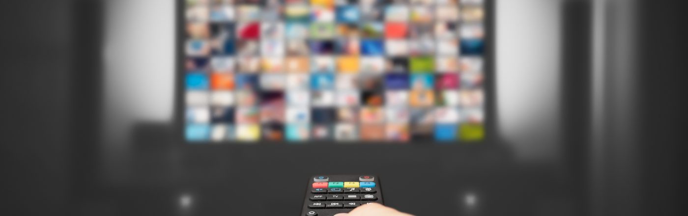 Auf dem Bild in dem Artikel zu den Reality-Shows 2026 ist eine Hand mit einer Fernbedienung im Vordergrund zu sehen, die auf einen großen, leicht verschwommenen Fernseher gerichtet ist. Auf dem Bildschirm erscheinen zahlreiche bunte Vorschaubilder.