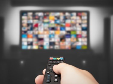 Auf dem Bild in dem Artikel zu den Reality-Shows 2026 ist eine Hand mit einer Fernbedienung im Vordergrund zu sehen, die auf einen großen, leicht verschwommenen Fernseher gerichtet ist. Auf dem Bildschirm erscheinen zahlreiche bunte Vorschaubilder.