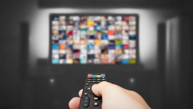 Auf dem Bild in dem Artikel zu den Reality-Shows 2026 ist eine Hand mit einer Fernbedienung im Vordergrund zu sehen, die auf einen großen, leicht verschwommenen Fernseher gerichtet ist. Auf dem Bildschirm erscheinen zahlreiche bunte Vorschaubilder.