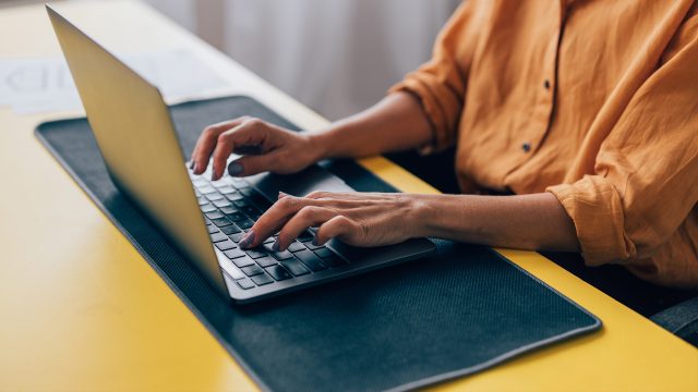 Auf dem Bild ist eine Person in einem orangefarbenen Hemd zu sehen, die konzentriert an einem Laptop arbeitet. Die Hände tippen aktiv auf der Tastatur, während der Arbeitsplatz mit einem gelben Schreibtisch und dunkler Unterlage modern und aufgeräumt wirkt. Das Bild symbolisiert produktives Arbeiten mit verschiedenen ChatGPT-Versionen in einer ruhigen Umgebung.