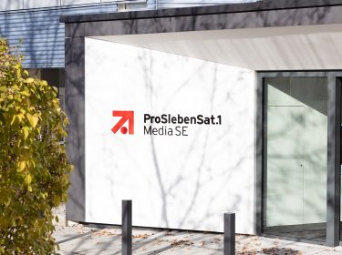 Auf dem Bild zur angekündigten Reality-Show The Hunt ProSieben ist der Eingangsbereich des ProSiebenSat.1 Media SE-Gebäudes bei Tageslicht zu sehen. Das moderne Gebäude zeigt das bekannte rot-schwarze Senderlogo auf weißem Hintergrund. Im Vordergrund befindet sich ein herbstlicher Baum mit gelbem Laub.