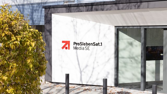 Auf dem Bild zur angekündigten Reality-Show The Hunt ProSieben ist der Eingangsbereich des ProSiebenSat.1 Media SE-Gebäudes bei Tageslicht zu sehen. Das moderne Gebäude zeigt das bekannte rot-schwarze Senderlogo auf weißem Hintergrund. Im Vordergrund befindet sich ein herbstlicher Baum mit gelbem Laub.