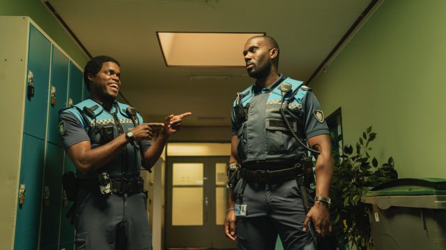 Bild aus Almost Cops: Zwei Hilfspolizisten in Uniform stehen sich in einer Umkleide gegenüber. Während der eine grinsend auf den anderen zeigt, blickt ihn dieser wütend an.