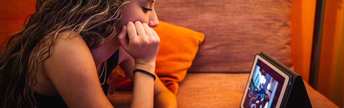 Ein junges Mädchen mit langen, gewellten Haaren liegt entspannt auf einem orangefarbenen Sofa und schaut konzentriert ein YouTube-Video auf einem Tablet. Die warme Beleuchtung mit kleinen Lichterketten im Hintergrund sorgt für eine gemütliche Stimmung. Die Szene steht exemplarisch dafür, ein Video ohne die automatischen YouTube-Übersetzungen anzuschauen.