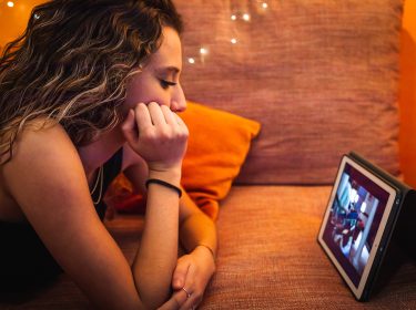 Ein junges Mädchen mit langen, gewellten Haaren liegt entspannt auf einem orangefarbenen Sofa und schaut konzentriert ein YouTube-Video auf einem Tablet. Die warme Beleuchtung mit kleinen Lichterketten im Hintergrund sorgt für eine gemütliche Stimmung. Die Szene steht exemplarisch dafür, ein Video ohne die automatischen YouTube-Übersetzungen anzuschauen.