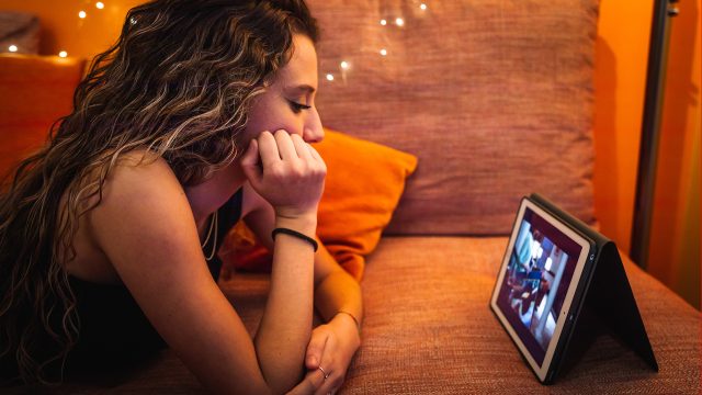 Ein junges Mädchen mit langen, gewellten Haaren liegt entspannt auf einem orangefarbenen Sofa und schaut konzentriert ein YouTube-Video auf einem Tablet. Die warme Beleuchtung mit kleinen Lichterketten im Hintergrund sorgt für eine gemütliche Stimmung. Die Szene steht exemplarisch dafür, ein Video ohne die automatischen YouTube-Übersetzungen anzuschauen.