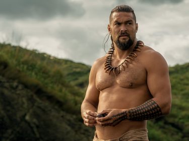 In dieser Szene aus der Chief of War-Serie ist Ka'iana, gespielt von Jason Momoa, zu sehen. Er steht oberkörperfrei in einer natürlichen, grünen Landschaft, trägt eine traditionelle Kette aus Zähnen und hat markante polynesische Tattoos am Arm und Bein. Der ernste Blick und die kraftvolle Körperhaltung verleihen der Szene aus der Chief of War-Serie eine entschlossene, kämpferische Stimmung.