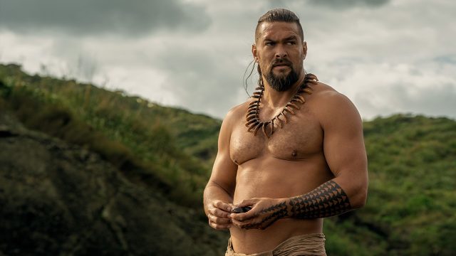 In dieser Szene aus der Chief of War-Serie ist Ka'iana, gespielt von Jason Momoa, zu sehen. Er steht oberkörperfrei in einer natürlichen, grünen Landschaft, trägt eine traditionelle Kette aus Zähnen und hat markante polynesische Tattoos am Arm und Bein. Der ernste Blick und die kraftvolle Körperhaltung verleihen der Szene aus der Chief of War-Serie eine entschlossene, kämpferische Stimmung.