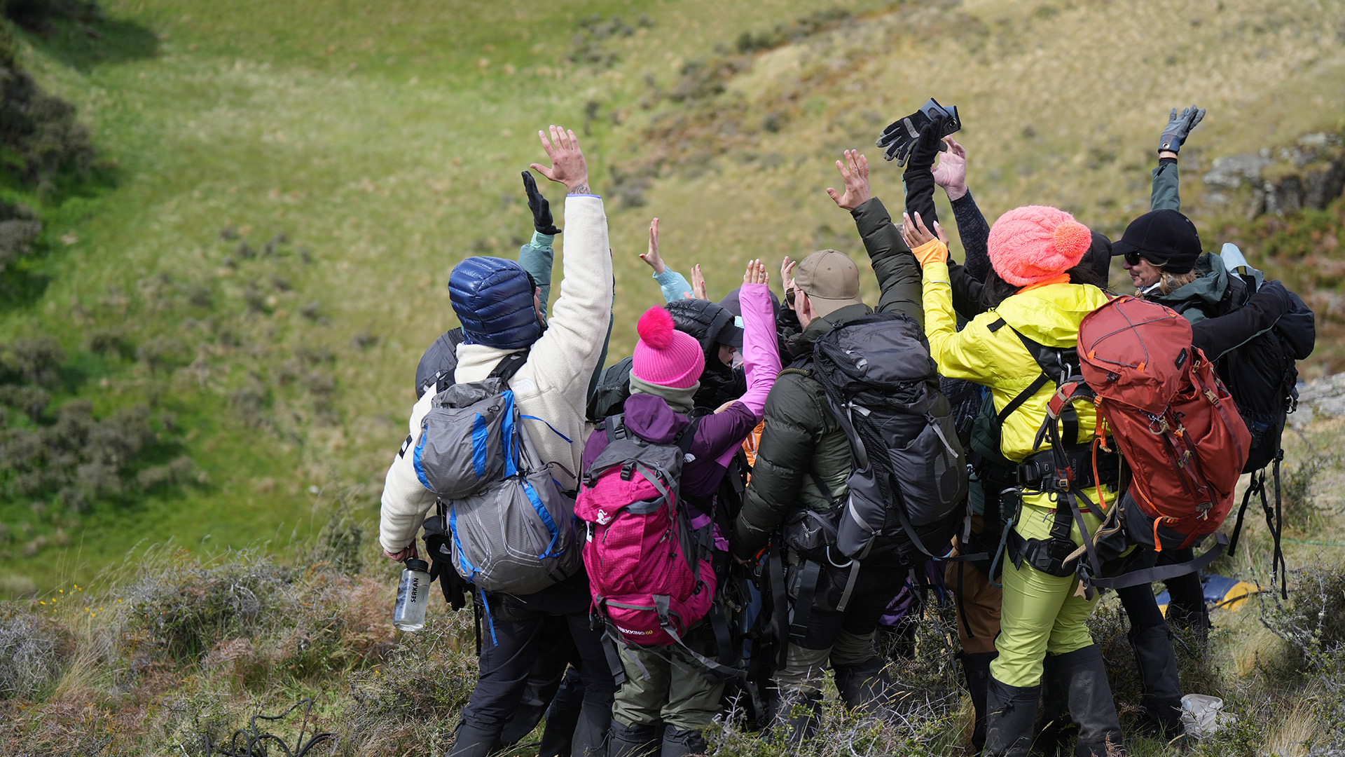 Auf dem Bild zur Reality-Show The Summit ist eine Gruppe von Teilnehmer*innen beim Wandern in einer bergigen Landschaft zu sehen. Alle tragen Outdoor-Kleidung, große Rucksäcke und heben im Kreis feierlich die Hände in die Höhe. Die Stimmung wirkt motiviert und gemeinschaftlich.