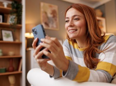 Rothaarige Frau liegt auf dem Sofa und schaut auf ihr iPhone